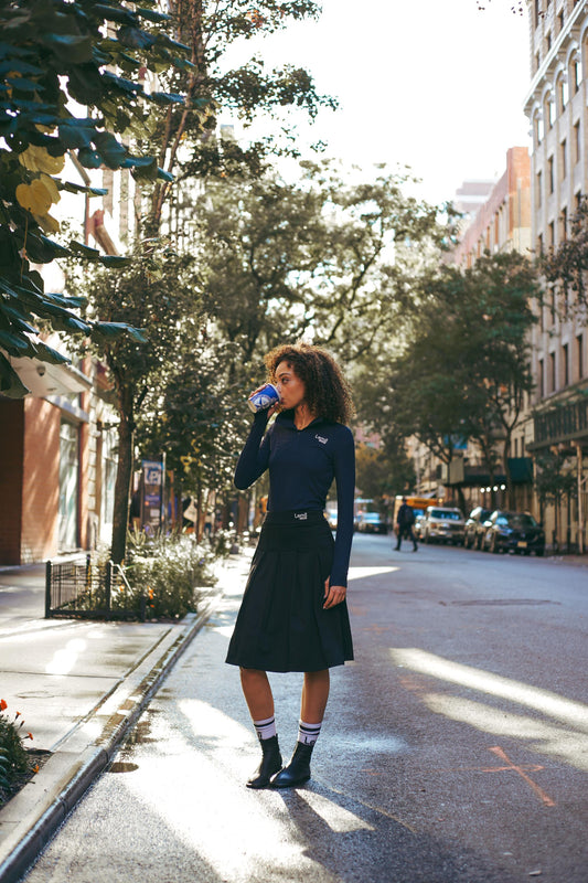 Lemii Label - Woman standing on a city street drinking from a coffee cup wearing navy athletic ribbed half up top, performance pleated skirt.