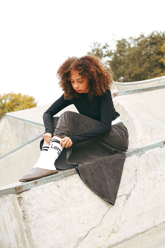Lemii Label - Person sitting on a concrete ledge wearing a black top and gray skirt with white socks.