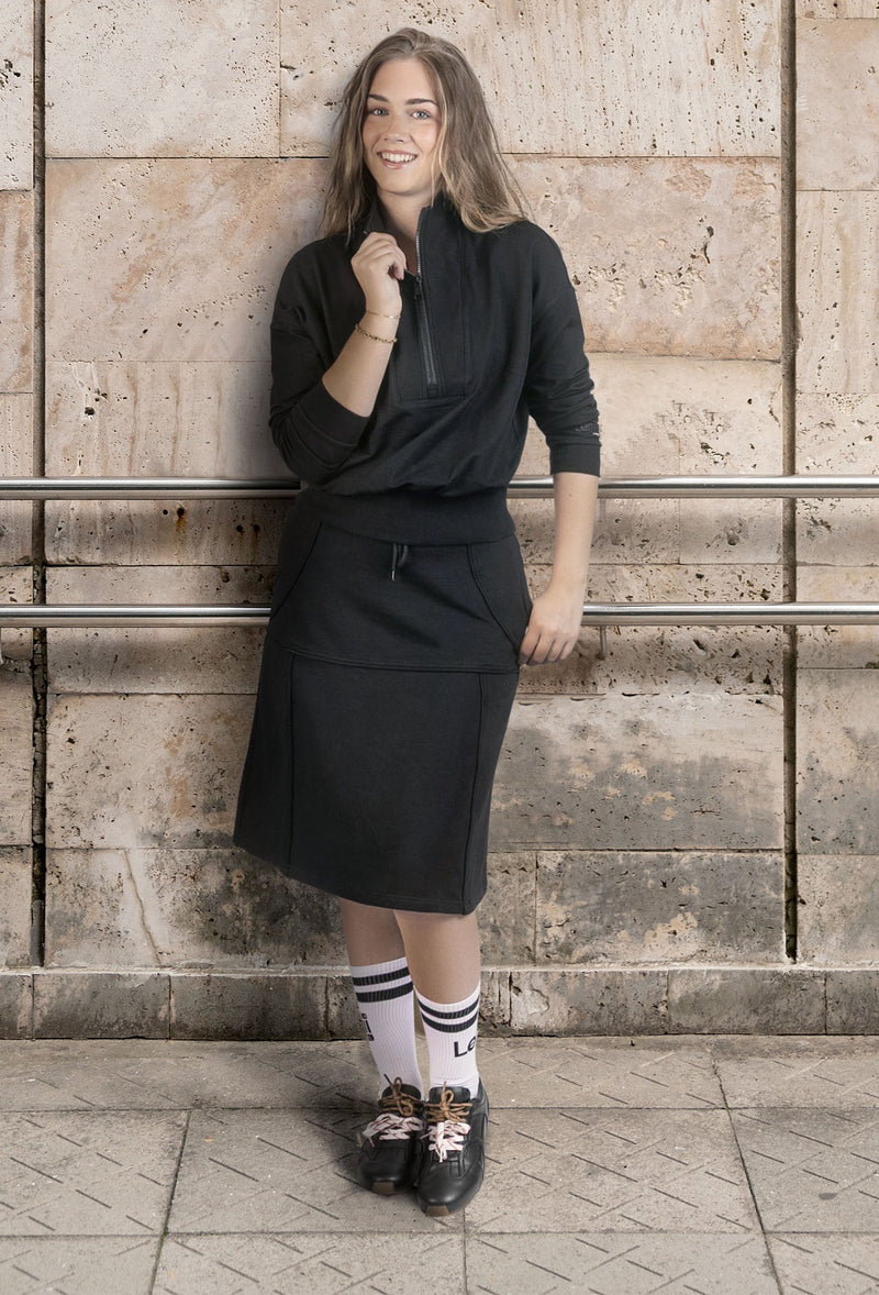 A woman wearing a black long-sleeve half-zip top and matching black skirt stands against a textured stone wall, wearing white socks and black shoes.