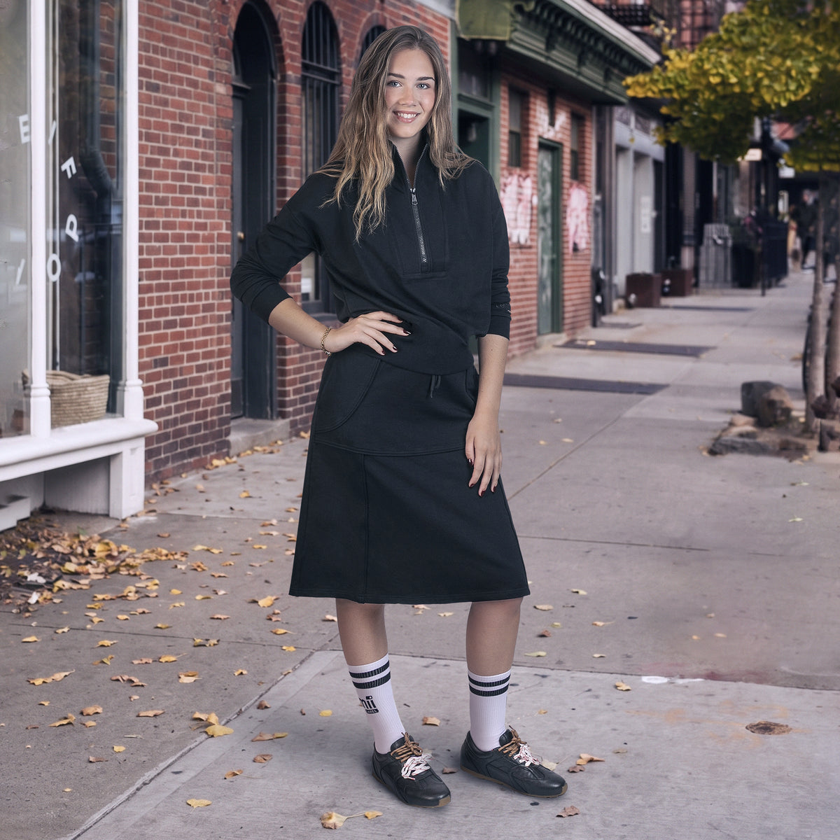 A woman stands on a city sidewalk wearing a black wrap-style midi dress with a half-zip neckline and a gathered waist. She is also wearing white knee-high socks with black stripes and black loafers with leopard print laces - lemii Label