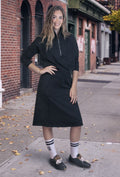 A woman modeling a black long-sleeve wrap-style midi dress with a half-zip neckline and side pockets, standing on a city sidewalk in front of brick buildings.