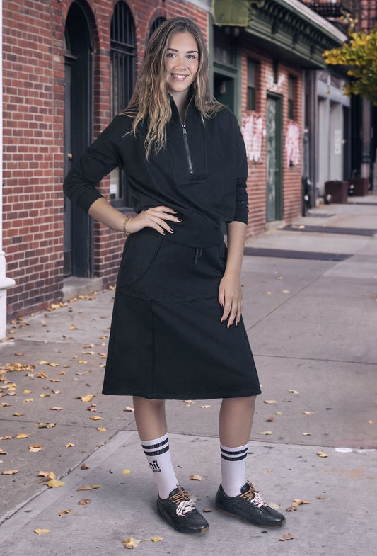 A woman modeling a black long-sleeve wrap-style midi dress with a half-zip neckline and side pockets, standing on a city sidewalk in front of brick buildings.