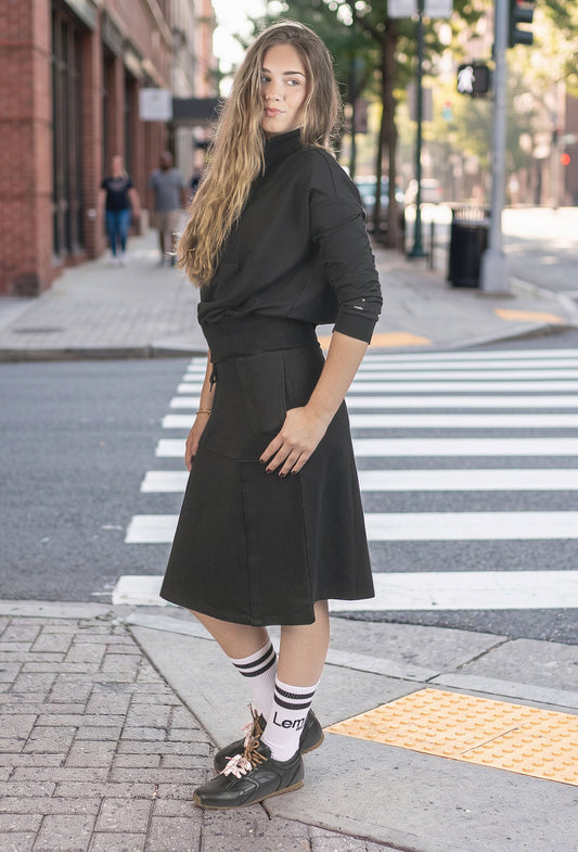 A woman stands on a city crosswalk wearing a black long-sleeve mock-neck top and a black A-line midi skirt, paired with white socks and dark black shoes.