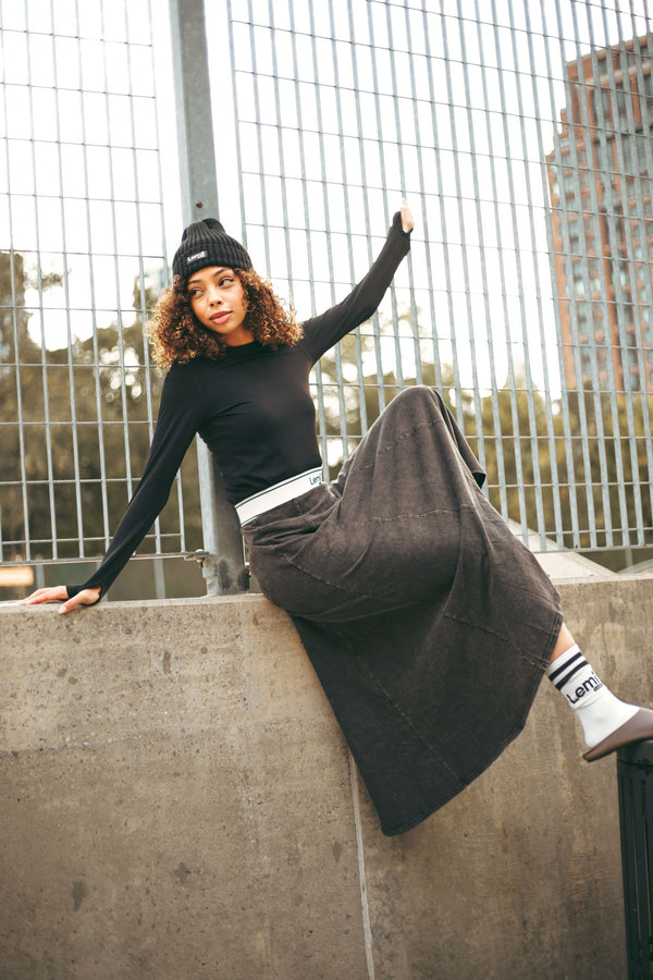 Lemii Label - Person sitting on a concrete ledge wearing a black athletic top and black wash skirt with a cityscape background.