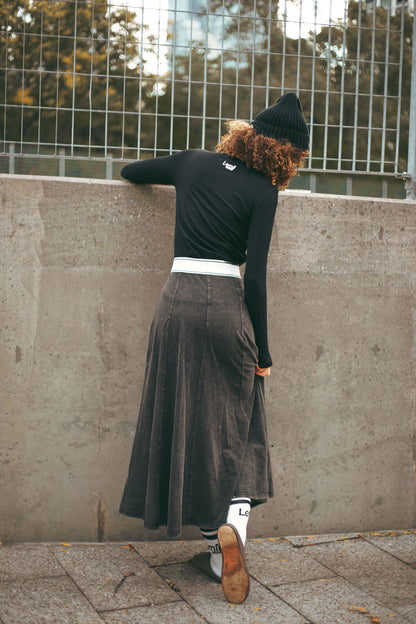 Person wearing a black sweater and gray skirt leaning against a wall.
