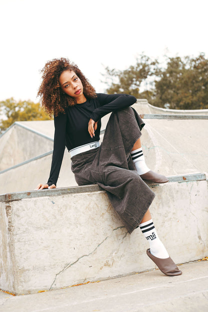 Person sitting on a concrete ledge wearing a black long-sleeve top, gray sweatskirt, and white socks with black stripes.