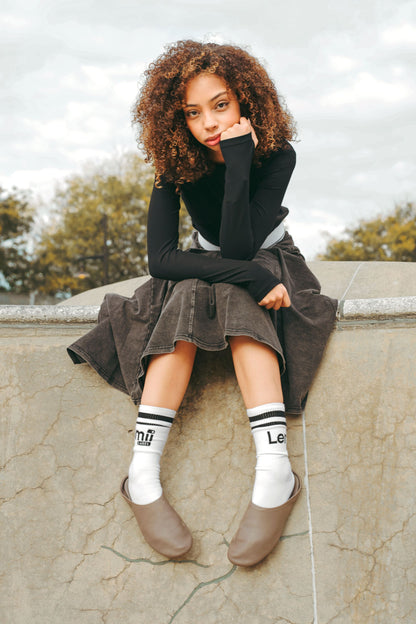 Lemmi Label - Woman sitting on a stone wall wearing a black athlitic top, black wash panel skirt, and white socks with black logo. 