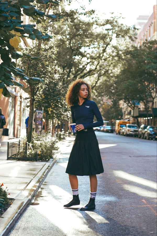 Lemii Label - Woman standing on a city street with trees and buildings in the background. Wearing black athletic skirt, navy athletic top. 