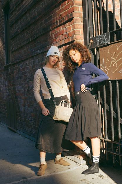 Two women standing against a brick wall, wearing stylish outfits with bags.