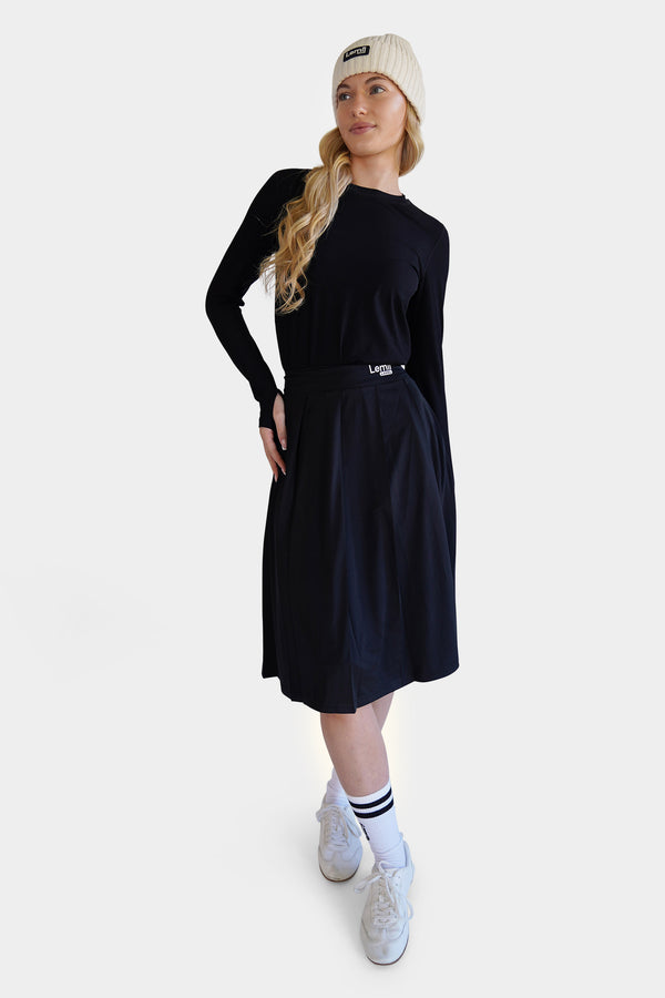 Woman wearing a black athletic pleated skirt, white sneakers, and a cream beanie on a white background - Lemii Label
