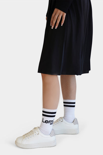 Person wearing a black dress, white sneakers, and socks with a brand logo on a white background