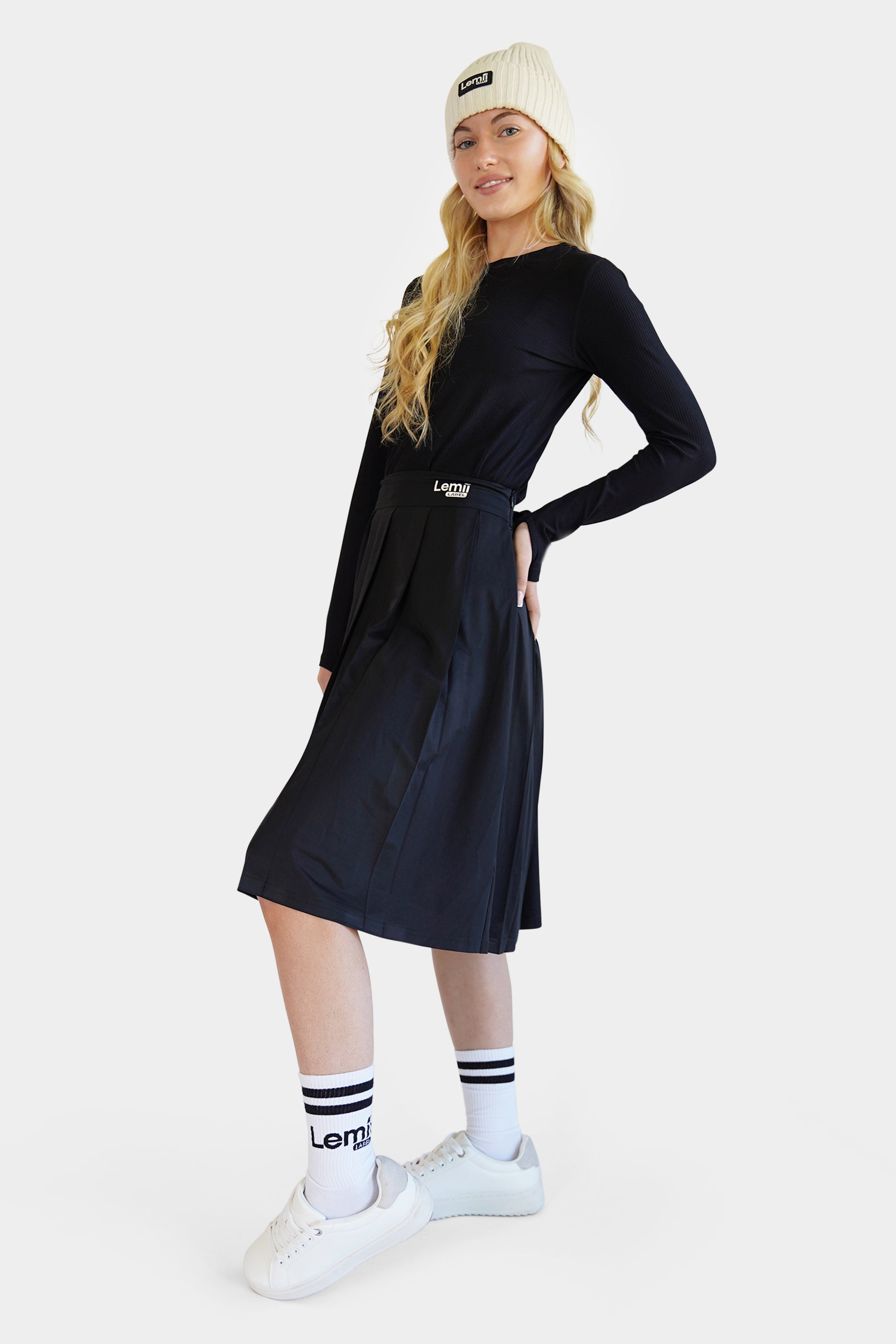 Lemii Label - Woman wearing a black athletic pleated skirt and top, white sneakers, and a cream beanie on a white background