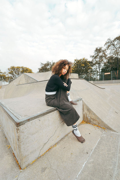 Person sitting on a concrete ledge at a skate park with trees and sky in the background wearing a grey skirt
