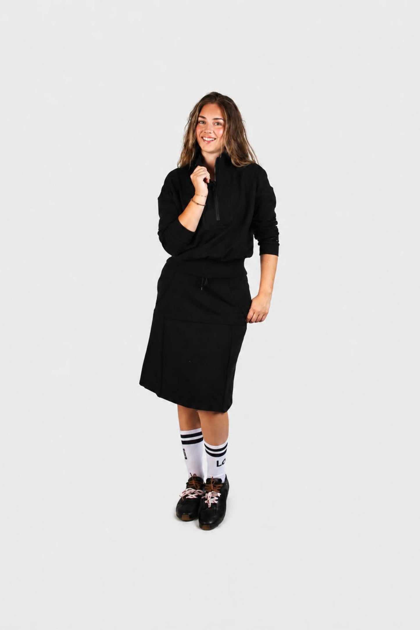 A young woman modeling a black long-sleeve knit dress with a drawstring waist, paired with white socks and black sneakers, against a solid light gray background  - Lemii Label