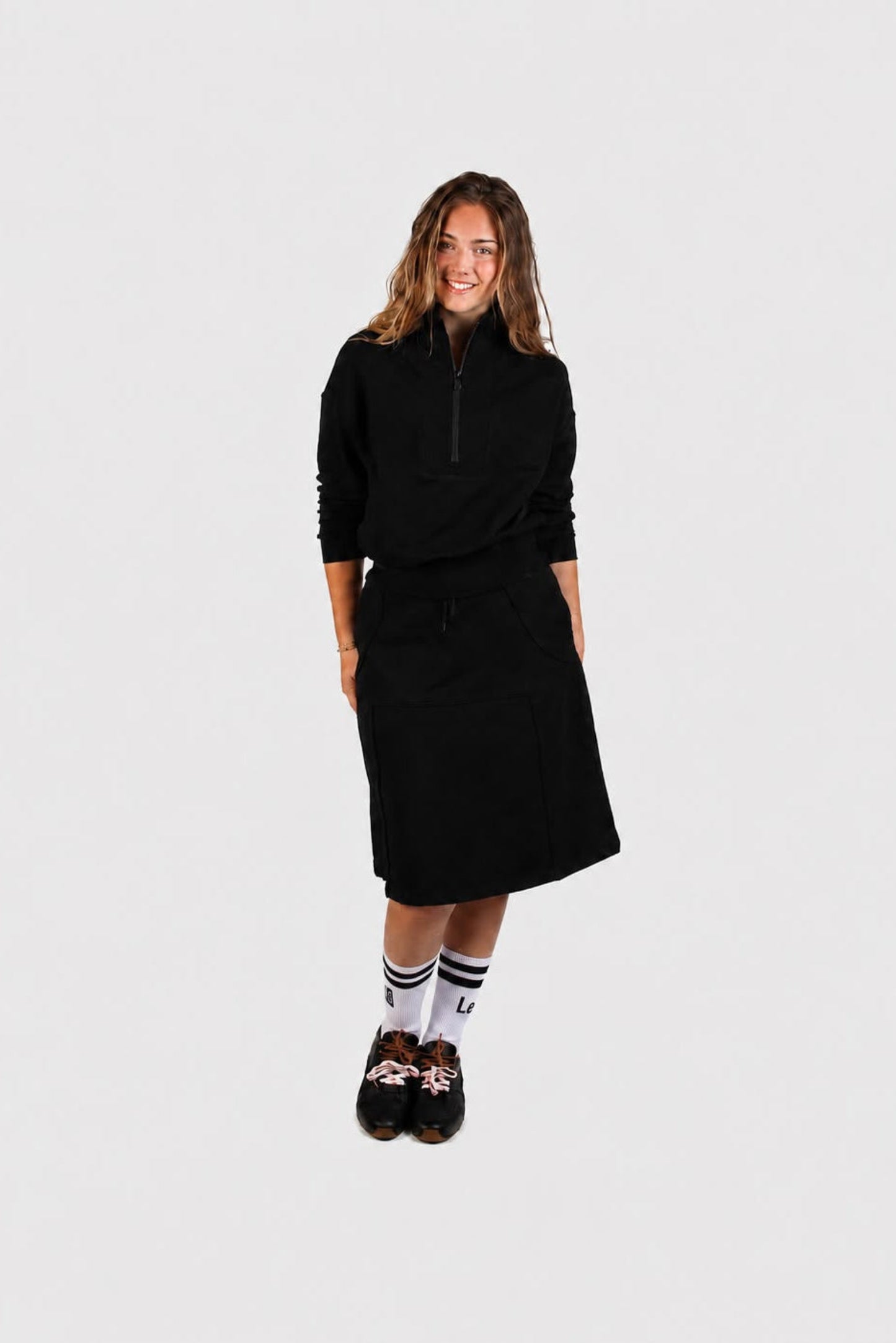 A woman modeling a black long-sleeve half-zip pullover and matching black midi skirt set, paired with white socks and black loafers, against a solid light gray background - Lemii Label