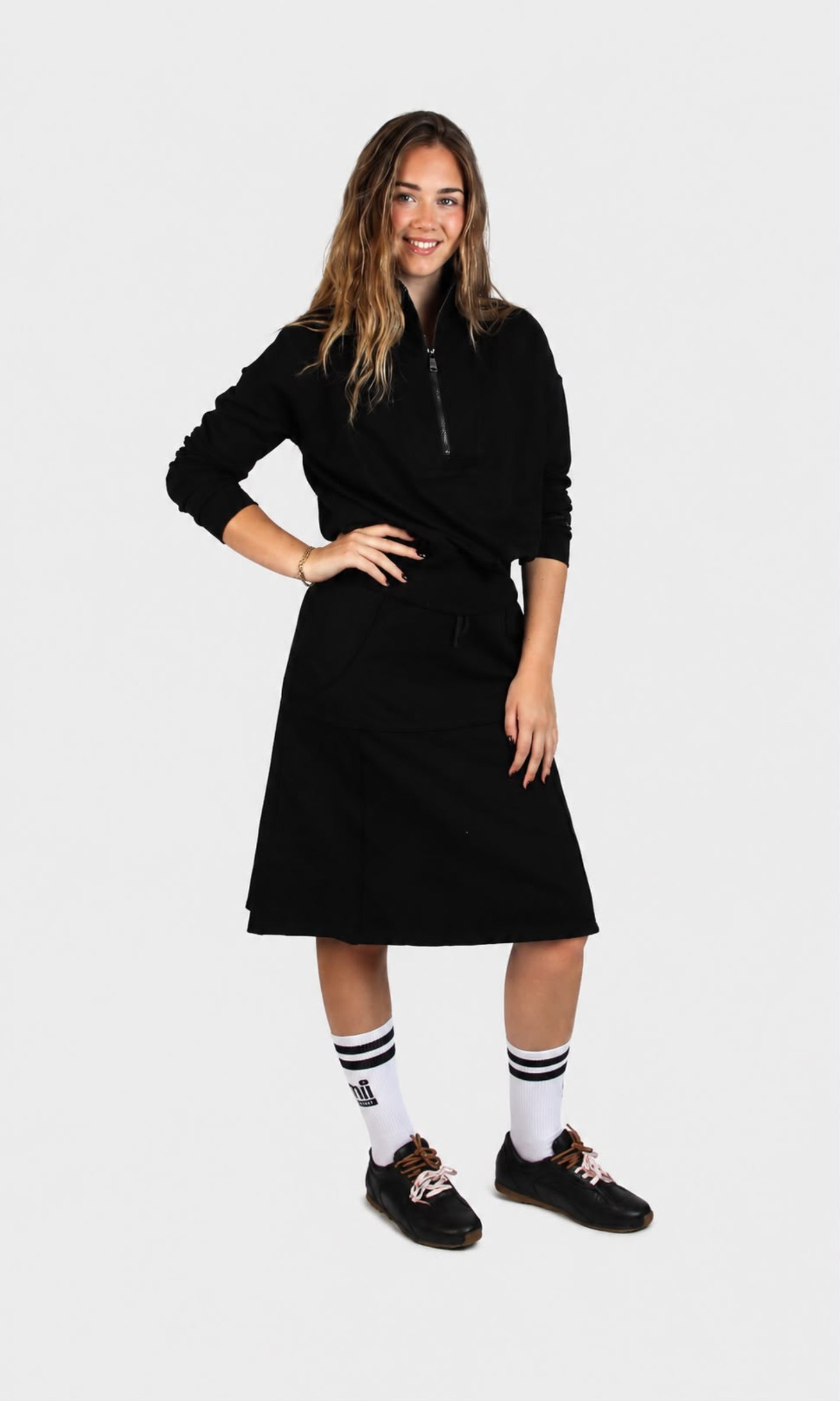 A woman modeling a black long-sleeve half-zip dress with a drawstring waist, paired with white socks and black shoes, against a light gray background - Lemii Label