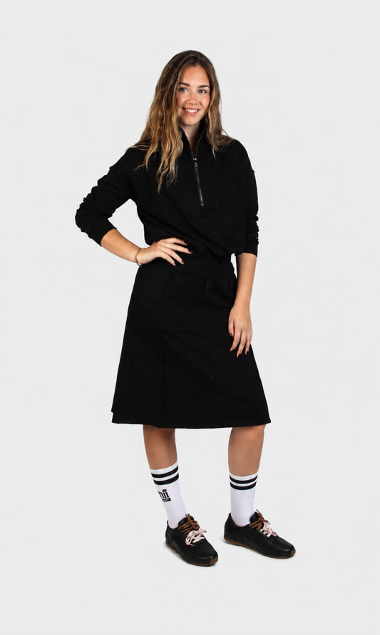 A woman modeling a black long-sleeve half-zip dress with a drawstring waist, paired with white socks and black shoes, against a light gray background - Lemii Label