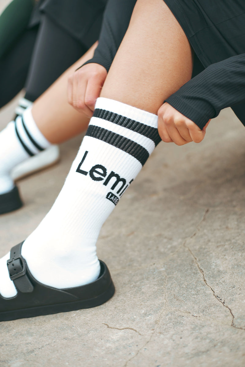 Person wearing white socks with black stripes and Lemii label branding, sitting on a concrete surface.