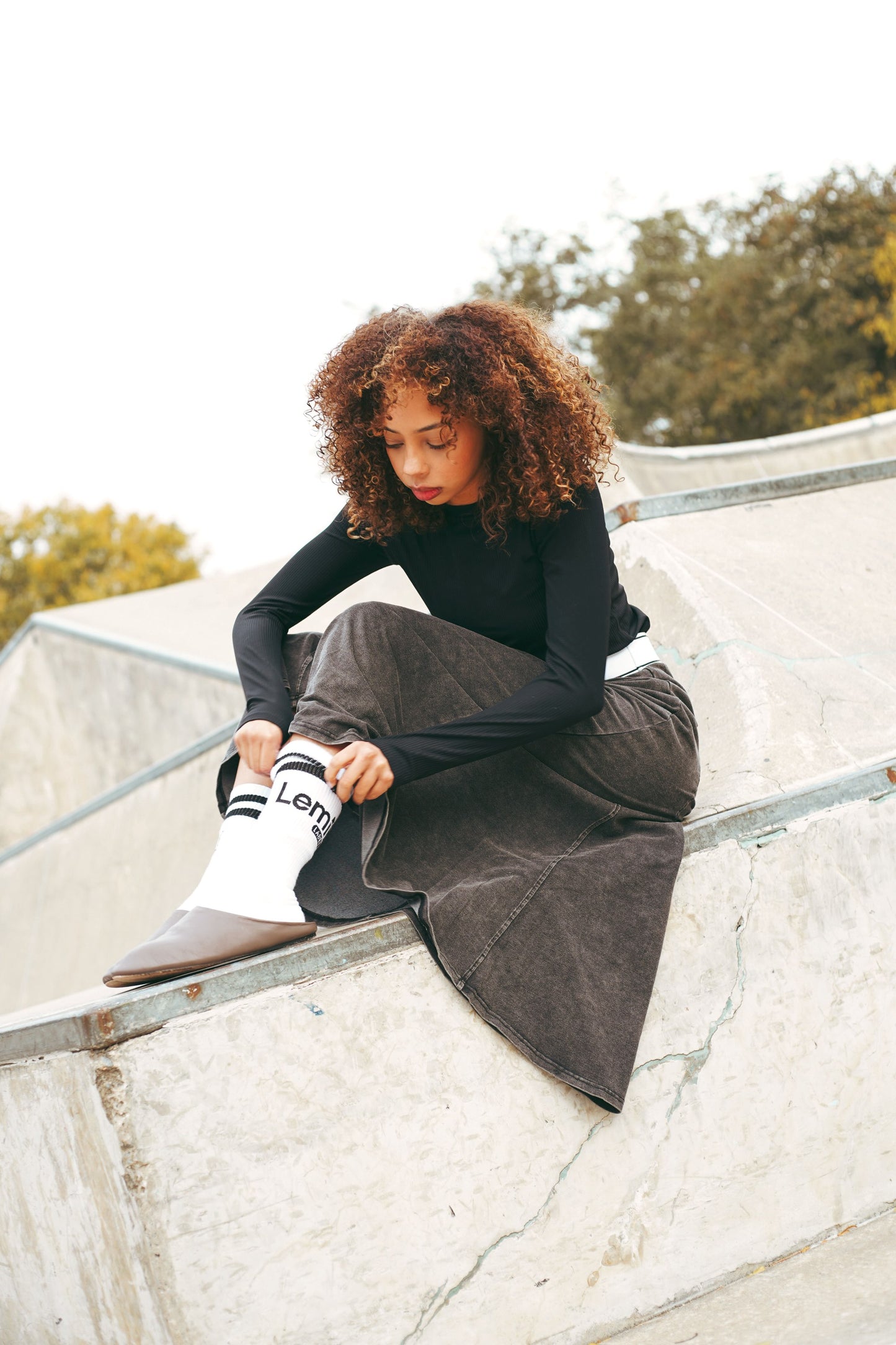 Lemii Label - Person sitting on a concrete ledge wearing a black top and gray skirt with white socks.