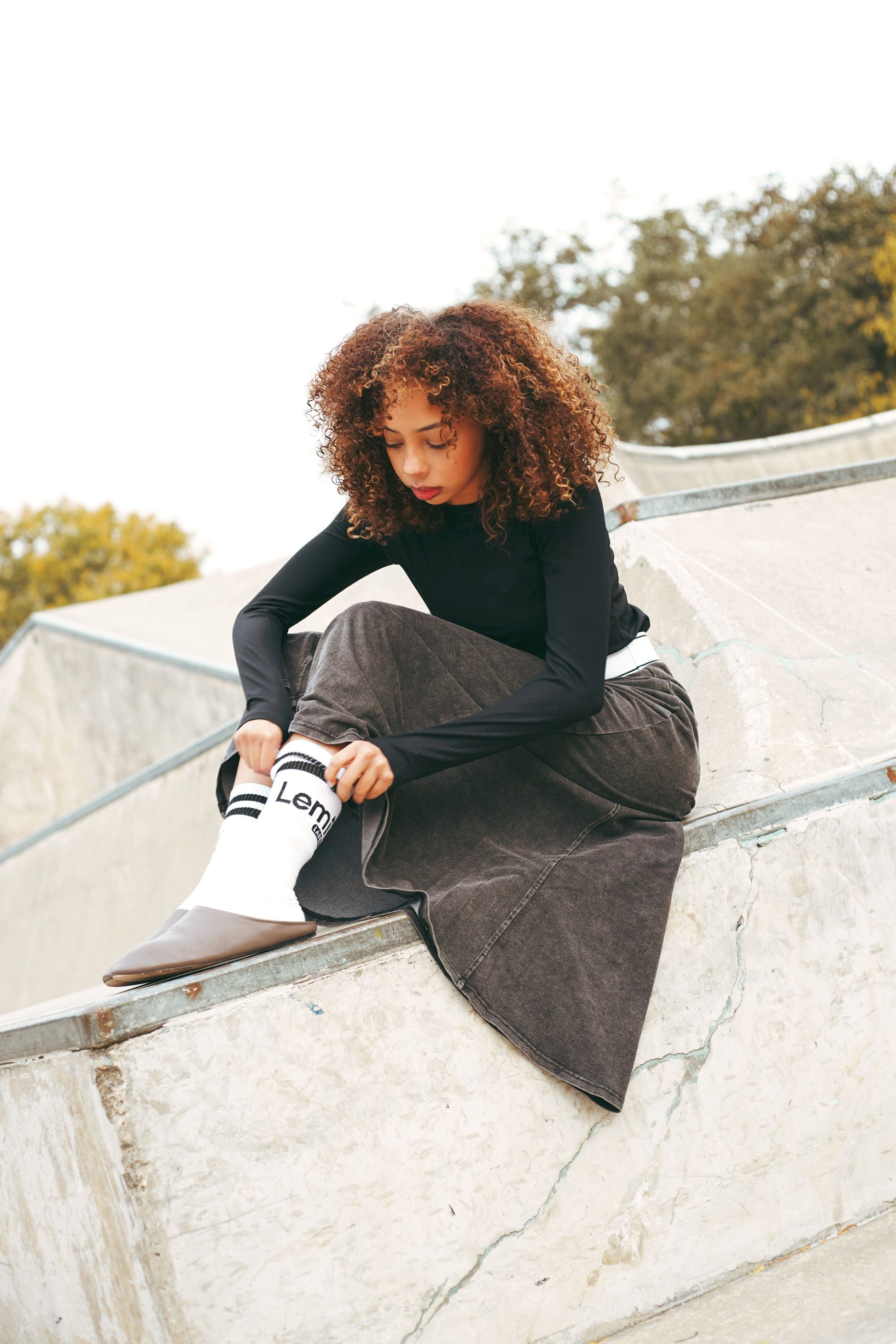 Lemii Label - Person sitting on a concrete ledge wearing a black top and gray skirt with white socks.